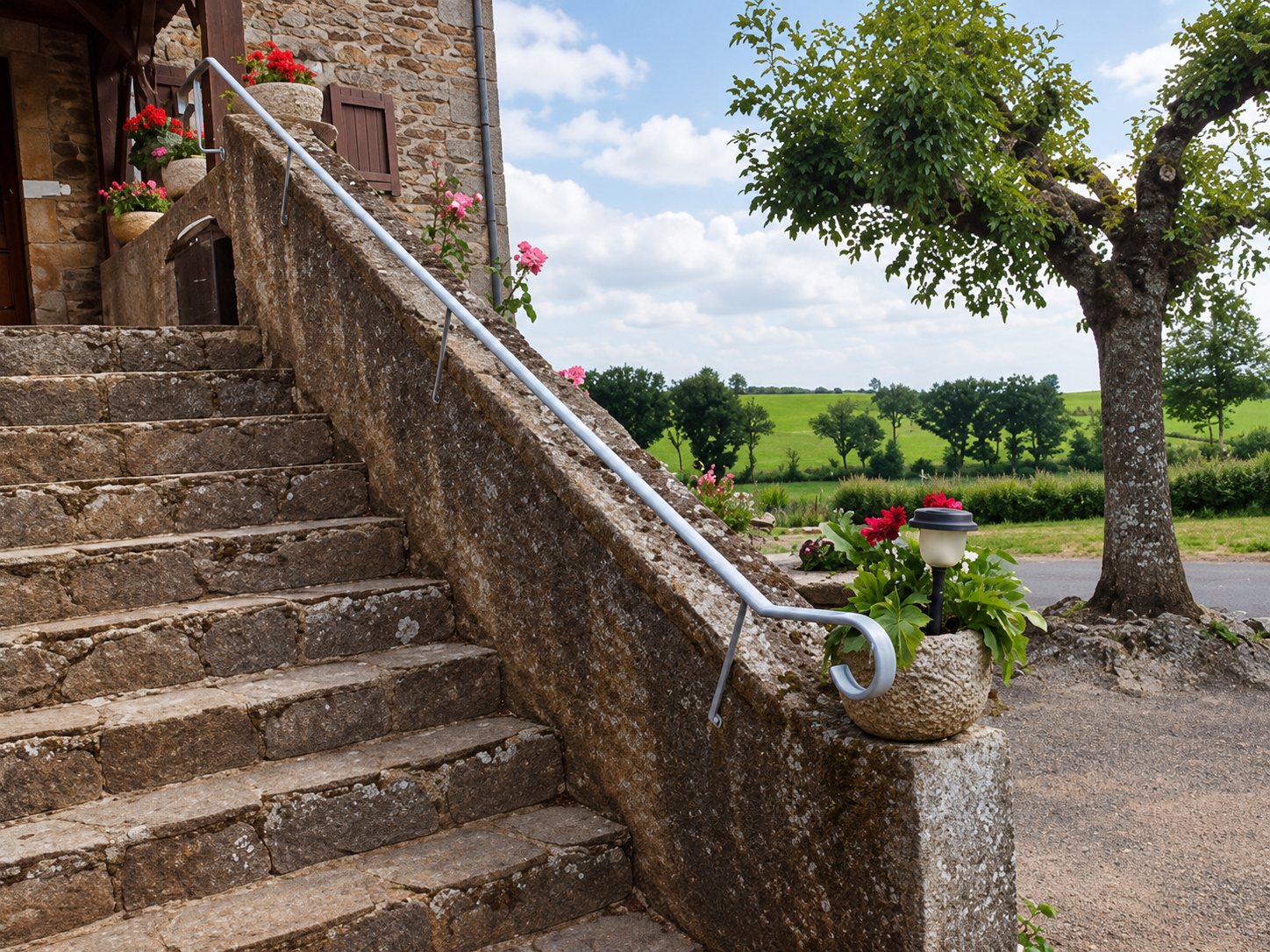 Escalier en pierre avec rampe métallique devant un paysage de campagne verdoyant