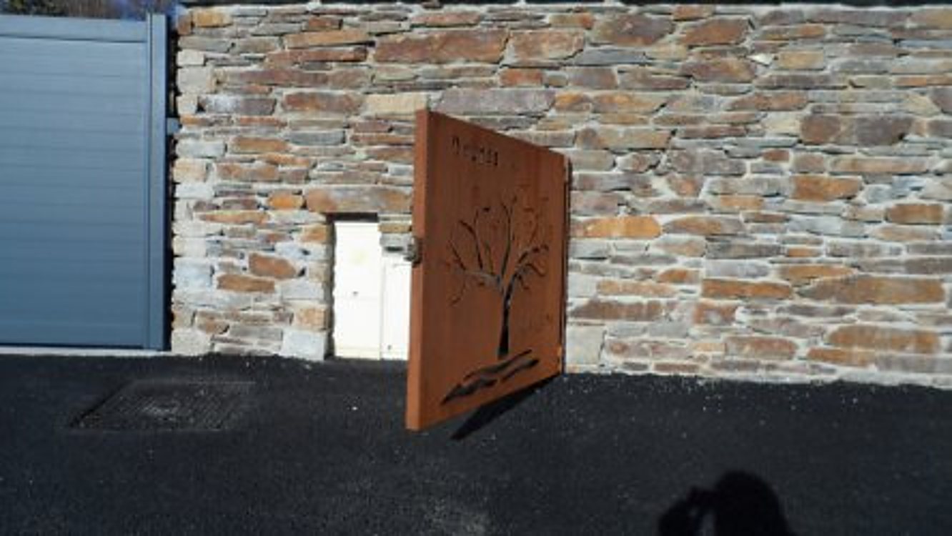 Portillon en acier corten orné d’un arbre devant un mur en pierre